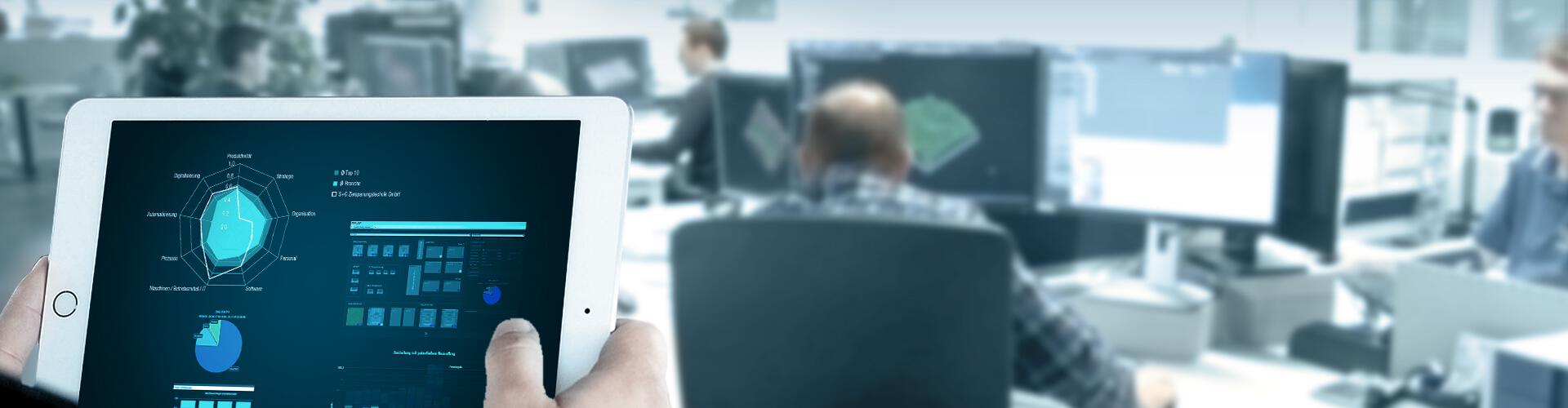 Hand mit Tablet, das Betriebsdaten aus der Fertigung verdichtet in Diagrammen anzeigt. Im Hintergrund ist ein CAM-Programmierbüro. 
