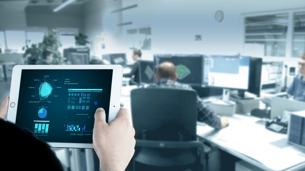 Hand mit Tablet, das Betriebsdaten aus der Fertigung verdichtet in Diagrammen anzeigt. Im Hintergrund ist ein CAM-Programmierbüro. 
